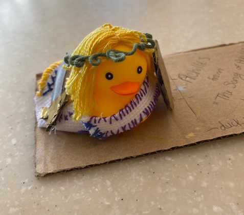 A small yellow rubber duck is dressed in a handmade embroidered toga and a vine crown. On its left side is a cardboard sword and on its right is a cardboard sheild.