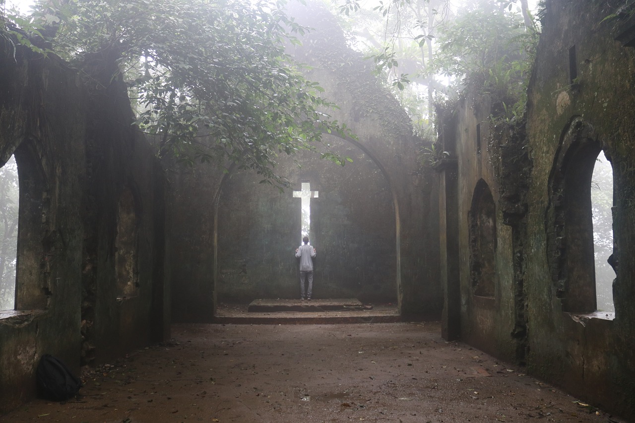 And inside view of an abandoned church or abbey. There is a hazy misty quality to the photo that makes it seem otherworldly. The roof of the building is long gone and there are vines and trees growing up and around the walls.  A person stands before a cross shaped window cut into the far wall.