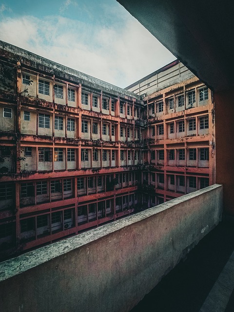 An open to the outdoors hallway looks out on a hundred small windows of an old abandoned hospital building. The building is a mix of beige, gray, and rusty red in colors.