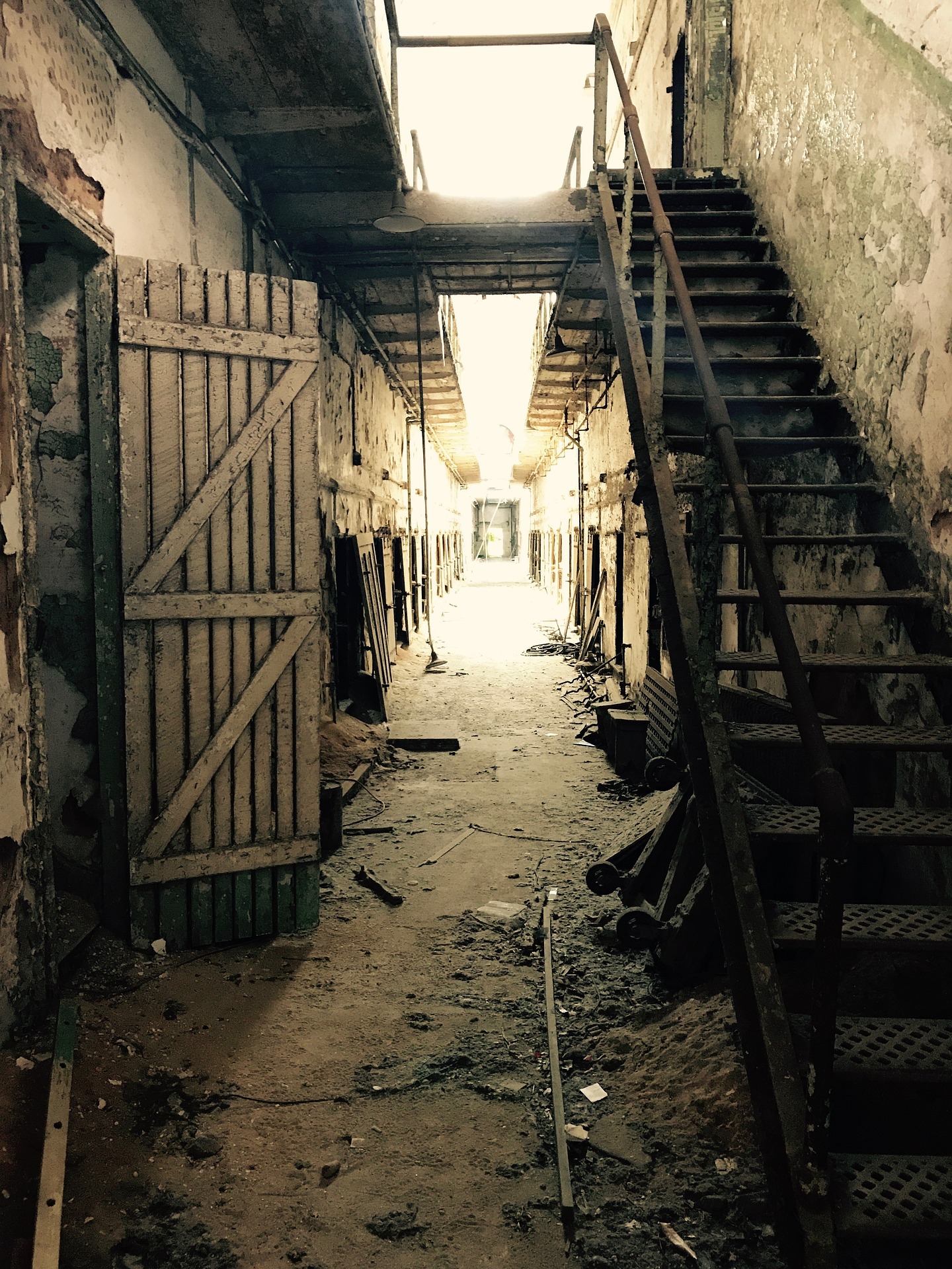 A dilapidated penitentiary hallway with a crumbling set of stairs to the right of the photo and rotting doors along the hallway to the left. 