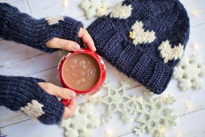Two gloved hands hold a red mug of hot chocolate next to a knitted hat and some decorative snowflakes.