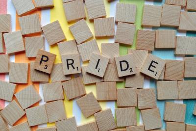 Over some rainbow colored stripes are a bunch of blank scrabble tiles with five tiles spelling out the word "Pride".