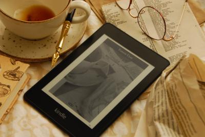 Kindle showing cover of book sourrounded by empty tea cup, glasses, amd papers