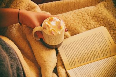 a person sits in a comfy looking spot with a blanket and a book across their lap and a hot chocolate in their hand.