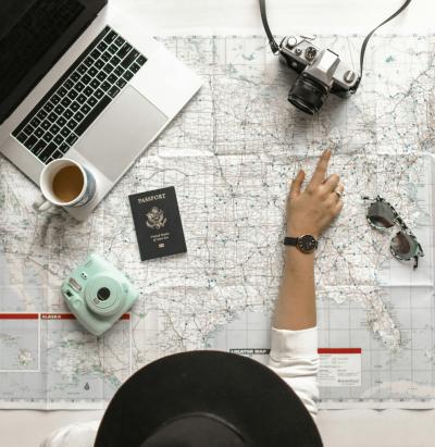 Photo of a hand pointing at a map, a computer, two cameras, a passport, and sunglasses.