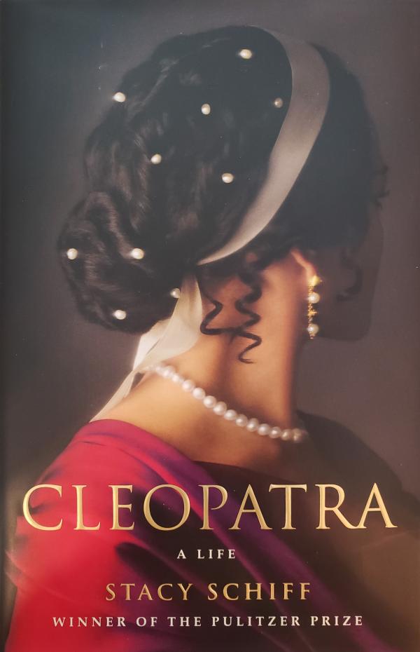A woman in a pinkish read dress with pearls around her neck, in her ears, and in her dark brown/black curly hair faces away from the reader. 