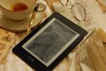 Kindle showing cover of book sourrounded by empty tea cup, glasses, amd papers