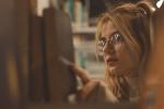 A young blonde woman with glasses reaches her hand forward to take a book off of a shelf. The camera view is looking through the shelf at the woman.