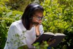 A woman sits among the foliage pensively reading a book. She has on a white t-shirt, glasses, and black headphones.